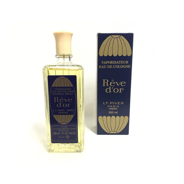 A bottle of Rève d'or Eau de Cologne by LT Piver, featuring a clear glass bottle filled with pale yellow liquid and a cream-colored cap, standing next to its navy blue and gold retail box on a white background.