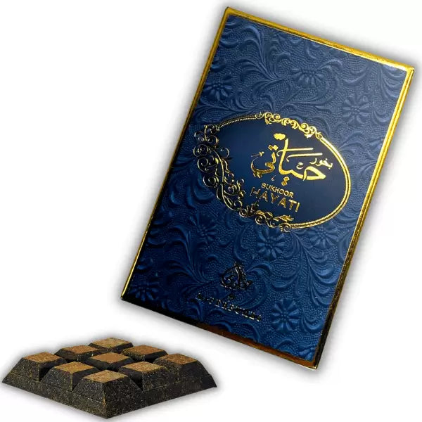A dark blue embossed chocolate box with gold foil detailing and Arabic calligraphy, shown alongside a small square sample of dark chocolate pieces on a white background.
