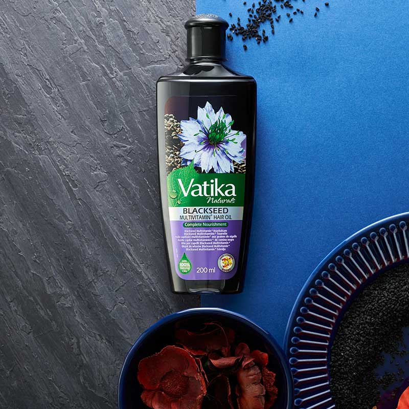 A bottle of Vatika Naturals Blackseed Mehitani Parthenol Oil (200ml) is displayed on a split gray and blue textured background, styled with dried black seeds and red rose petals in bowls.