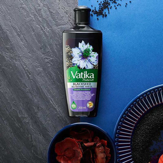 A bottle of Vatika Naturals Blackseed Mehitani Parthenol Oil (200ml) is displayed on a split gray and blue textured background, styled with dried black seeds and red rose petals in bowls.
