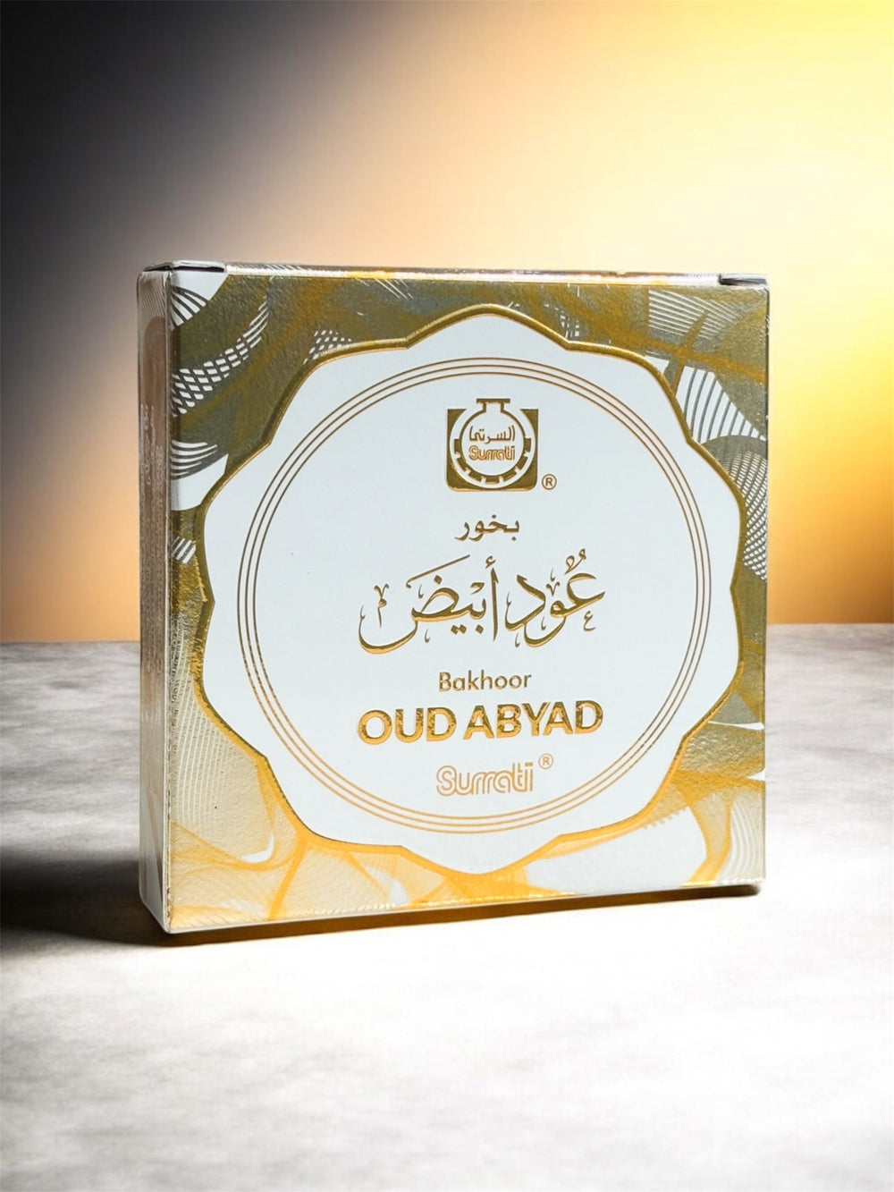A square product box for Oud Abiyad incense by Sureti, featuring a white central label with gold Arabic and English text, surrounded by a gold and gray patterned border. The box is photographed on a light gray surface against a warm yellow gradient background.