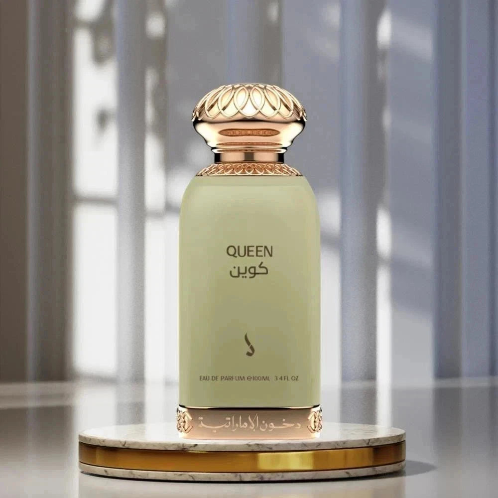 A perfume bottle labeled QUEEN in English and Arabic, with a pale sage green body and ornate gold-toned cap and base, displayed on a round marble-and-gold pedestal against a softly blurred interior background.Queen Perfume 100ml EDP Dkhoon Emirates
