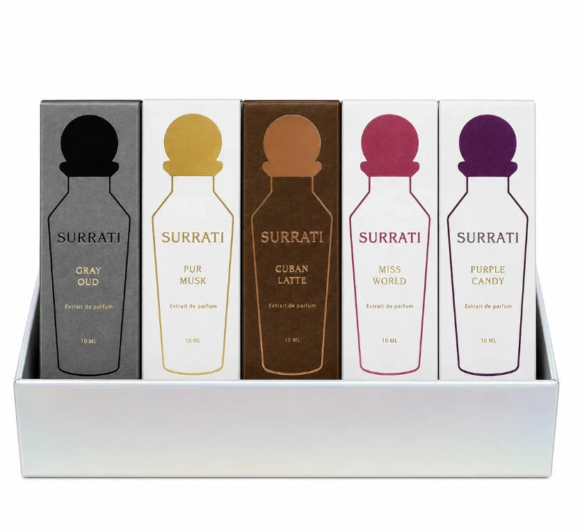 Five Surrati perfume bottles in a white display tray, each with its own colored cap and label: Gray Oud in dark gray, Pure Musk in white with gold, Cuban Latte in brown with gold, Miss World in white with pink, and Purple Candy in white with purple.