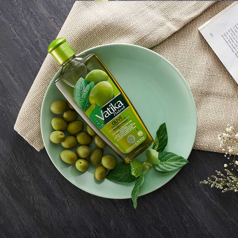 Vatika olive oil bottle on a plate with green olives and leaves on a dark surface.