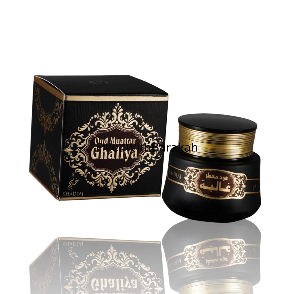 A black cosmetic jar with gold lettering next to its matching black and gold box, labeled 'Oud Muattar Ghaliya' by the brand Al-Rihla, on a white background with a reflection below.