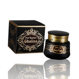 A black cosmetic jar with gold lettering next to its matching black and gold box, labeled 'Oud Muattar Ghaliya' by the brand Al-Rihla, on a white background with a reflection below.
