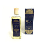 A bottle of Rève d'or Eau de Cologne by LT Piver, featuring a clear glass bottle filled with pale yellow liquid and a cream-colored cap, standing next to its navy blue and gold retail box on a white background.
