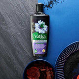A bottle of Vatika Naturals Blackseed Mehitani Parthenol Oil (200ml) is displayed on a split gray and blue textured background, styled with dried black seeds and red rose petals in bowls.
