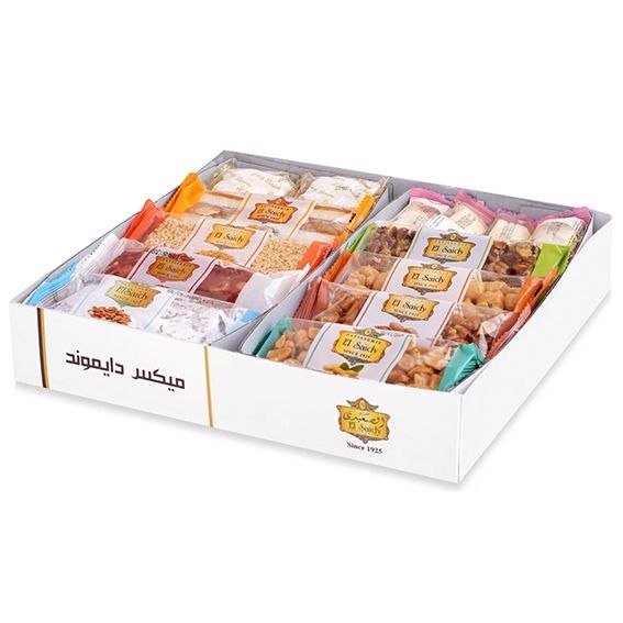 A white gift box filled with assorted packaged snacks and sweets, photographed on a clean white background. The box has Arabic text on the side and a small logo near the front right corner.