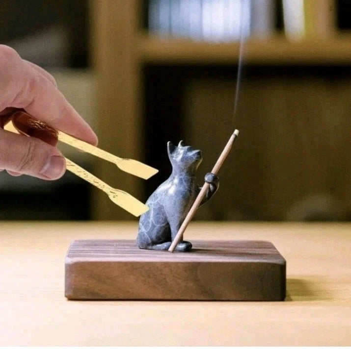 A small dark gray cat figurine holding a burning incense stick on a rectangular wooden base, with a hand using gold tongs to hold the incense holder in a softly blurred indoor setting.