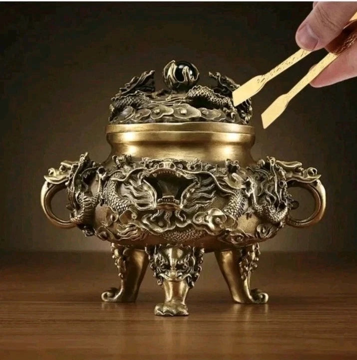 An ornate bronze-colored dragon incense burner with three legs and side handles, shown on a wooden surface with a hand holding tweezers near the lid.