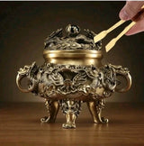 An ornate bronze-colored dragon incense burner with three legs and side handles, shown on a wooden surface with a hand holding tweezers near the lid.