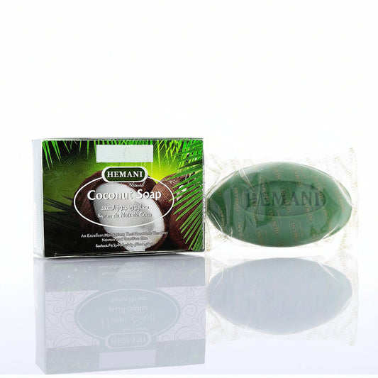 Hemani Coconut Soap shown with its retail box on a reflective white surface. The soap bar is green and wrapped in clear foil, and the box features coconut and palm leaf graphics with the Hemani brand name.