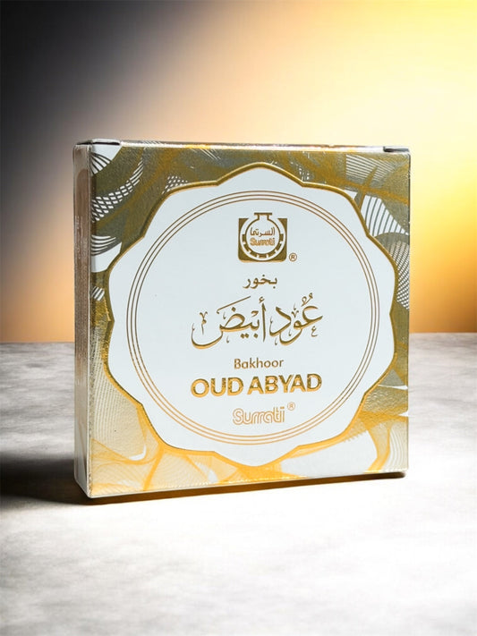 A square product box for Oud Abiyad incense by Sureti, featuring a white central label with gold Arabic and English text, surrounded by a gold and gray patterned border. The box is photographed on a light gray surface against a warm yellow gradient background.