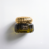 A small dark amber glass jar with a shiny gold ribbed lid, featuring Arabic and English text on the front, set against a clean white background with a soft  shadow. Mamoul Kuwaiti Bakhoor 60g – Gulf Incense