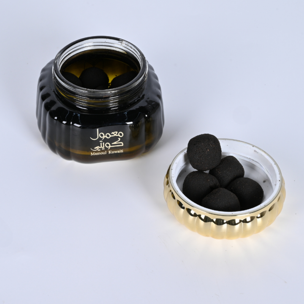 An open dark amber glass jar with Arabic text on the front, shown next to its gold-rimmed lid. Several round black compressed puffs are visible inside the jar and resting on the white interior of the lid. Mamoul Kuwaiti Bakhoor 60g – Gulf Incense