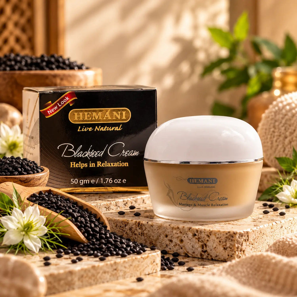 Hemani Blackseed Cream 50g jar and packaging box displayed on a stone slab with black seeds, flowers, and green leaves in a warm, natural setting.