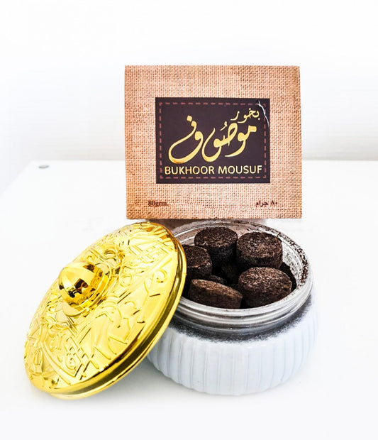 An open white glass jar filled with dark brown chocolate bougie confections, shown with a shiny gold decorative lid leaning in front and a textured brown product box behind it on a white surface.Mousuf Bakhoor 80g – Ard Al Zaafaran