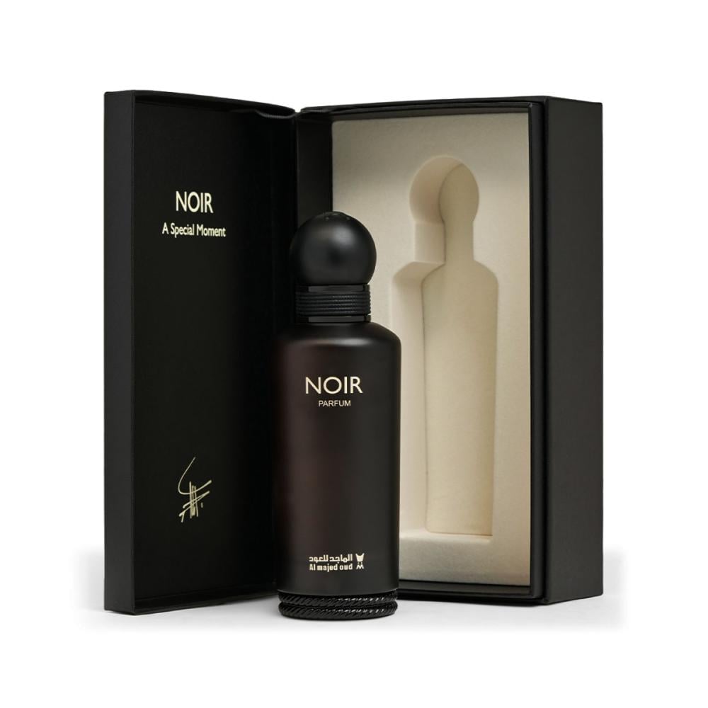 A dark brown glass perfume bottle labeled 'NOIR PARFUM' stands in front of an open black presentation box with a cream-colored insert. The box lid is printed with 'NOIR A Special Moment' and a small signature-like mark.Noir – Woody Fruity EDP 150ml – Al Majed Oud