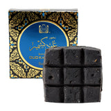 A square dark charcoal-colored soap bar with a 3x3 grid texture, shown in front of its ornate black and blue packaging box with gold details. The box features a prominent blue circular label with gold lettering.