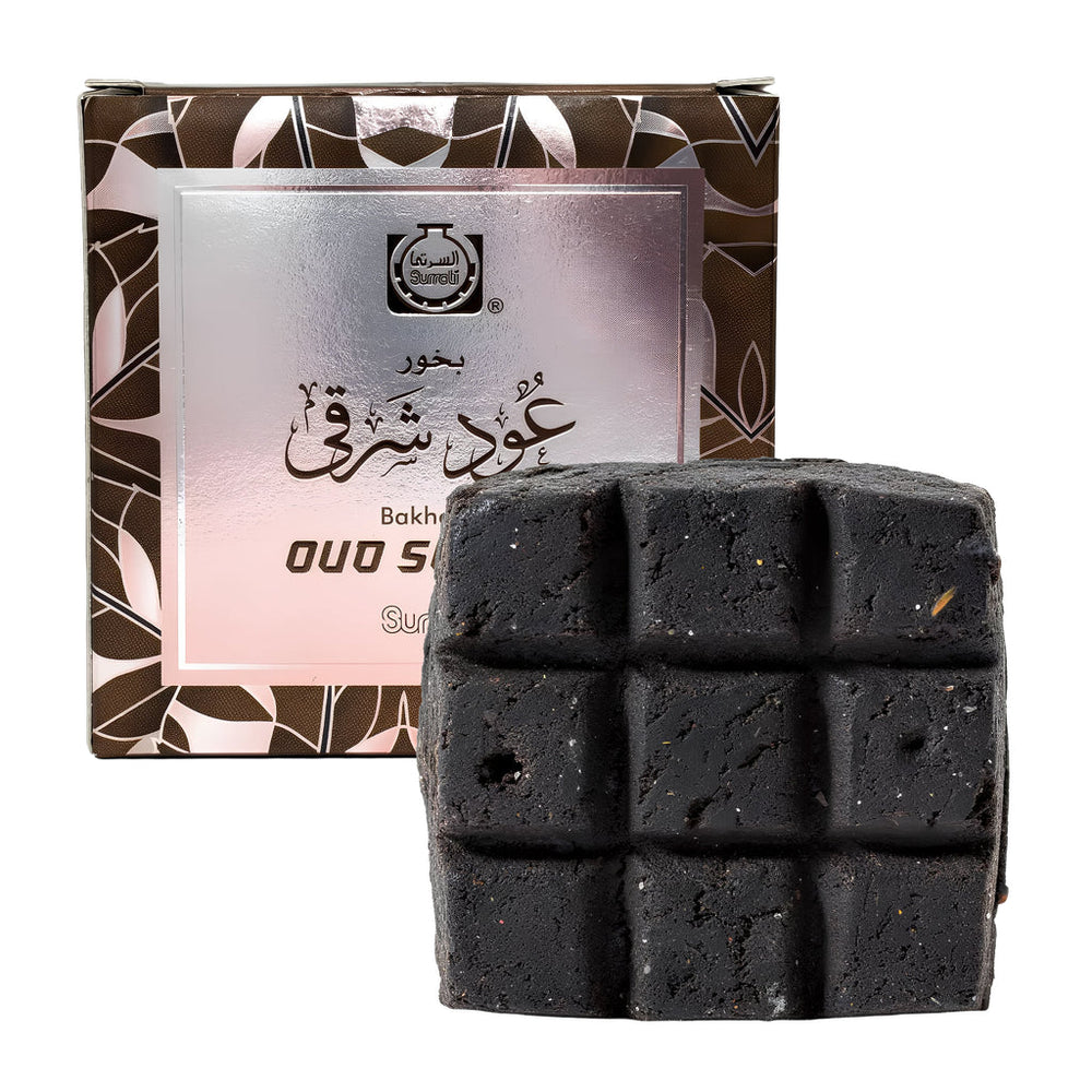 A dark charcoal-colored baked soap bar with a grid pattern, shown in front of its brown and silver patterned packaging box on a white background.