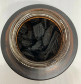 A top-down view of a clear glass jar filled with dark charcoal or carbon chunks, set against a light off-white background.Oud Muattar Munawwara Bakhoor 35g – Khadlaj