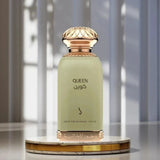 A perfume bottle labeled QUEEN in English and Arabic, with a pale sage green body and ornate gold-toned cap and base, displayed on a round marble-and-gold pedestal against a softly blurred interior background.Queen Perfume 100ml EDP Dkhoon Emirates
