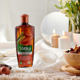 A bottle of Vatika Tindamri Moroccan Argan Moisturizing Hair Oil stands on a white textured blanket in a styled setting with lit candles, a bowl of dried botanicals, and soft floral decor in the background.