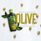A bottle of Vatika Multivitamin Enriched Olive Hair Oil is displayed on a textured white background, surrounded by scattered olive seeds and green leaves. The bottle is dark with a green cap and features a label with green olives.