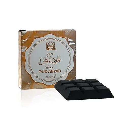 Black rectangular bukhoor oud abyad incense block with grid pattern next to ornate gold and white surrati packaging
