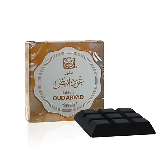 Black rectangular bukhoor oud abyad incense block with grid pattern next to ornate gold and white surrati packaging