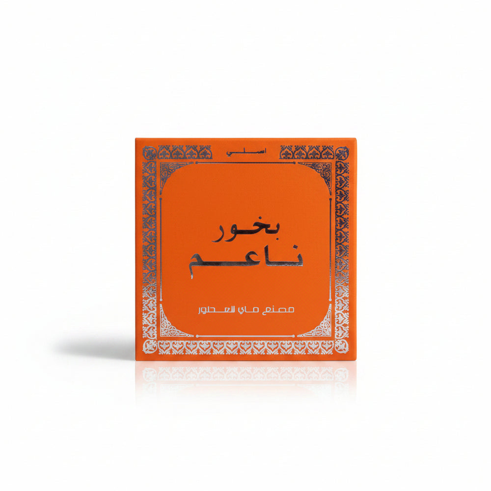 An orange square product box with Arabic text and a decorative white border, placed on a beige woven fabric background.