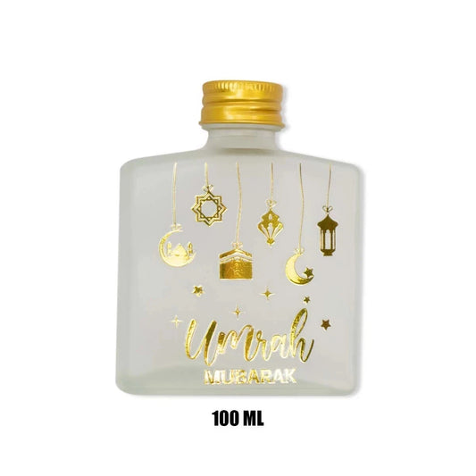 A 100 ml frosted glass bottle with a gold cap, decorated with gold foil illustrations of hanging lanterns, crescent moons, and stars, and the words 'Umrah MUGARAK' printed in gold. The bottle is shown against a plain white background with a '100 ML' text overlay below it.